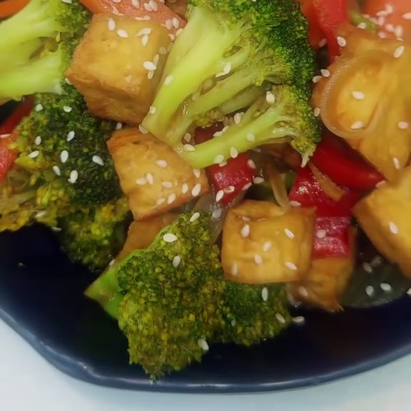 Broccoli florets, cubes of golden fried tofu, red bell pepper and carrot slices in a glossy brown sauce, scattered with white sesame seeds in a dark navy bowl at Pinoy Tambayan
