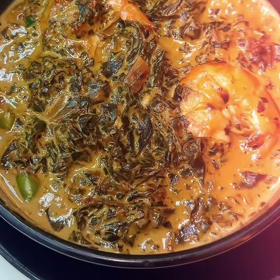 Close-up of laing, shredded taro leaves in an orange-tinted coconut sauce with a whole shrimp and a green chilli, in a dark pan at Pinoy Tambayan