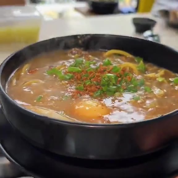 A full black bowl of lomi on a kitchen counter, thick brown broth topped with spring onions, toasted garlic and an egg, with yellow noodles around the rim at Pinoy Tambayan
