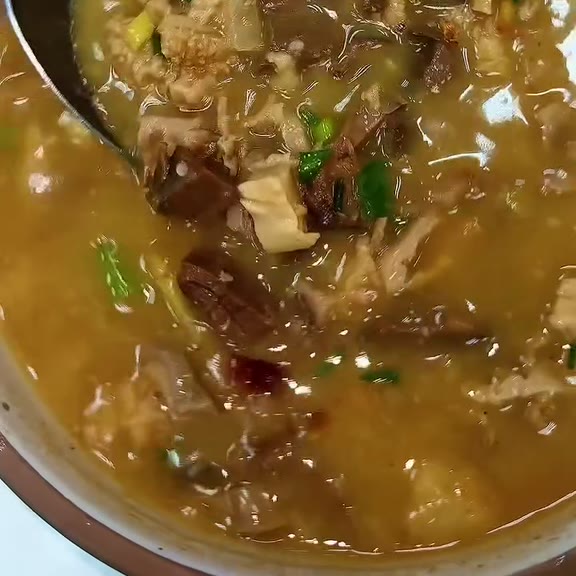 Close-up of papaitan, a brown beef offal soup with sliced tripe, chunks of beef and chopped spring onion in thick broth, served in a white bowl with a metal spoon resting in it at Pinoy Tambayan