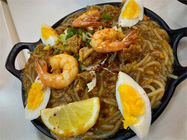 Sizzling pancit palabok topped with prawns, quartered boiled eggs, crispy chicharon, spring onion and a lemon wedge on a black cast iron plate