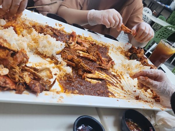 Gloved hands sharing a boodle fight platter of chilli crab, sweetcorn and rice spread across a long white tray
