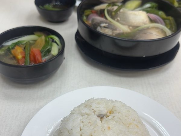 Sinigang na isda being ladled from a black pot at the table, with a plate of steamed rice in the foreground and a bowl of the sour broth