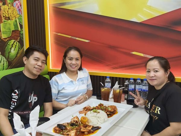 Three adult diners smiling at the camera behind a long platter of crab, prawns and fish in red sauce with corn and rice, in front of the Philippine flag mural