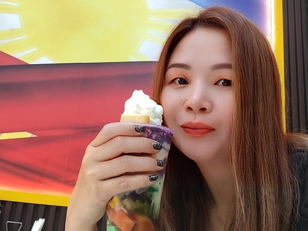 A woman holding up a tall cup of halo halo topped with whipped cream and ube, seated in front of the Philippine flag mural