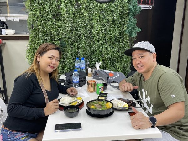 Two adult diners posing at a table set with a bulalo pot and two silog plates of rice and fried egg, under the restaurant's green foliage wall