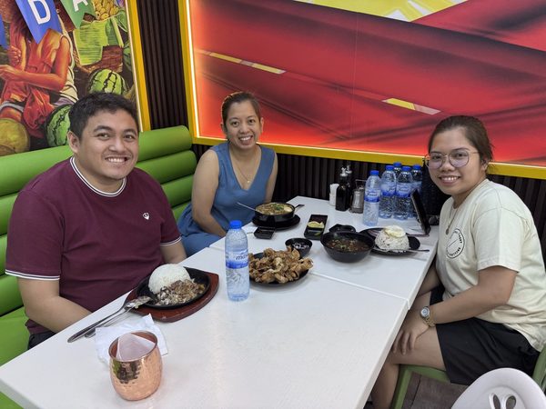 Three adult diners smiling at the camera at a table with sizzling sisig and rice, a plate of chicharon, bulalo bowls and a silog plate