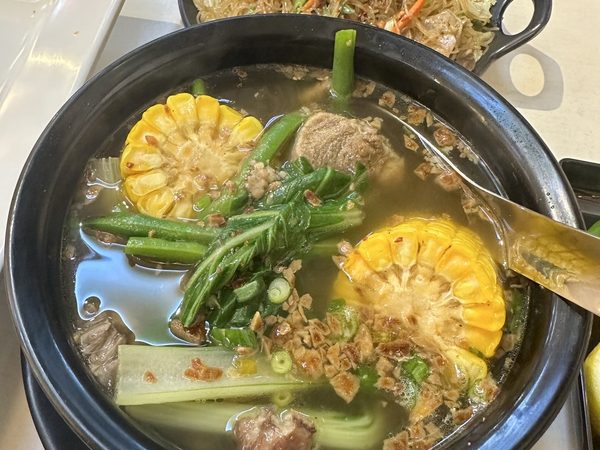 Bulalo beef shank soup with corn, okra, string beans and bok choy in a black pot, with a dish of pancit bihon behind it