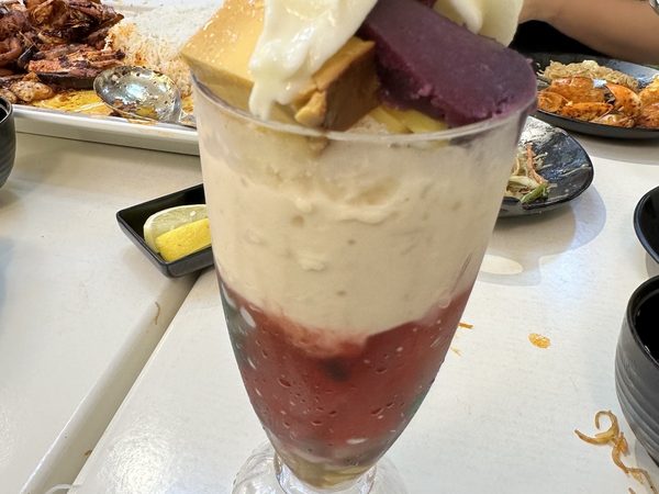 A tall glass of halo halo layered with leche flan, ube and whipped cream, on a table set with seafood platters