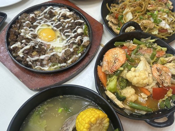 Shared table spread of sizzling sisig topped with a raw egg, pancit canton, bulalo with sweetcorn, chopsuey with prawns and steamed rice