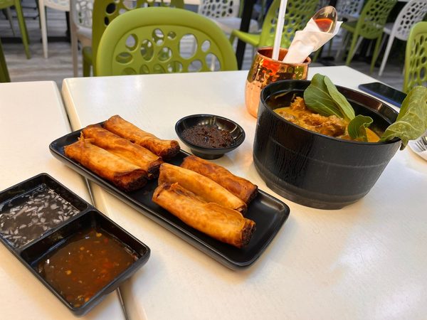 Two rows of crispy lumpia on a black plate with dipping sauces and a bowl of kare kare with bok choy alongside