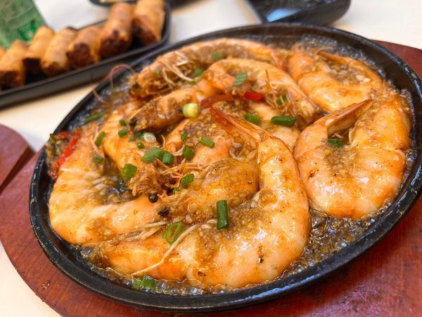Sizzling garlic butter prawns in a hot skillet with a plate of lumpia in the background