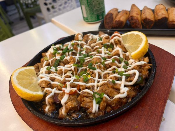 Sizzling chicken sisig drizzled with mayonnaise and topped with spring onion, with a tray of lumpia behind it