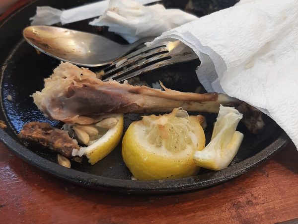 A finished sizzling plate with a pork bone, roasted garlic, squeezed lemon and used cutlery on a wooden board