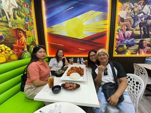 Four adult diners posing at a table set with sweet and sour prawns, steamed rice and glazed chicken wings, in front of the restaurant's Philippine flag mural