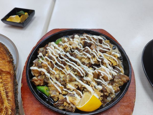 Sizzling beef sisig with mayonnaise drizzle and a lemon wedge, served with fried fish and a dish of pickled chilli and lemon