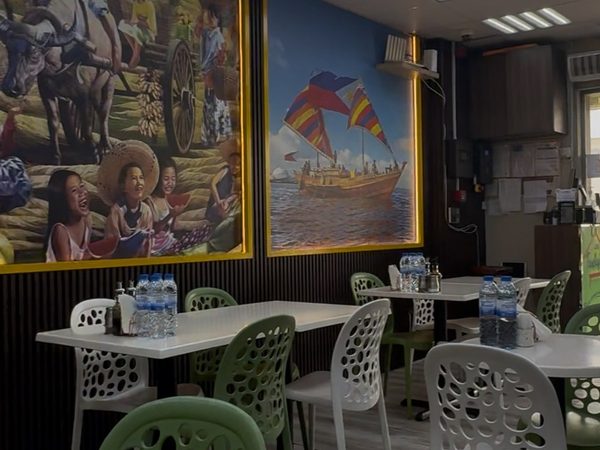 Empty dining room with white tables, green and white patterned chairs and large framed Filipino rural-scene paintings on a slatted wall.