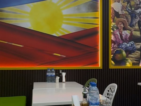 Corner of the dining area with a large Philippine flag painting and a harvest-scene mural above a round table set with condiments and bottled water.