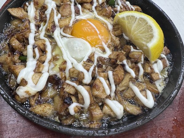 Sizzling chicken sisig on a hot plate with a raw egg yolk in the centre, mayonnaise drizzle and a lemon wedge.