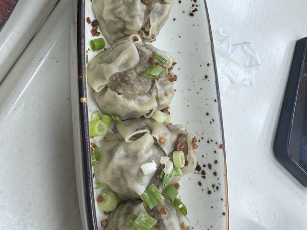 Four steamed pork siomai dumplings topped with spring onion and fried garlic on a long white plate, with a phone lying face-up at the edge of the table.