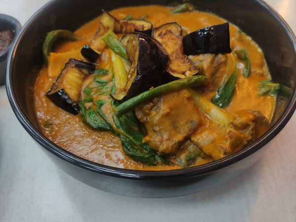 Bowl of kare kare, meat and vegetables in thick peanut sauce with fried eggplant, string beans and pechay, with a side of bagoong.