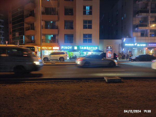 Night view of the illuminated Pinoy Tambayan restaurant sign and shopfront seen from across a busy road, with traffic passing in front.