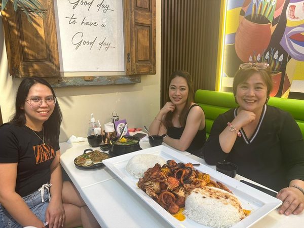 Three adult women smile at the camera in a booth behind a long seafood boodle platter with crab, squid rings, corn and mounds of rice.