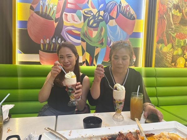Two adult women on a green banquette holding tall glasses of halo halo topped with ube soft serve and leche flan.