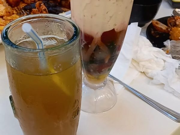Glass of halo halo with ube soft serve, leche flan and mixed jellies beside a tall iced tea, with a seafood platter on the table behind.