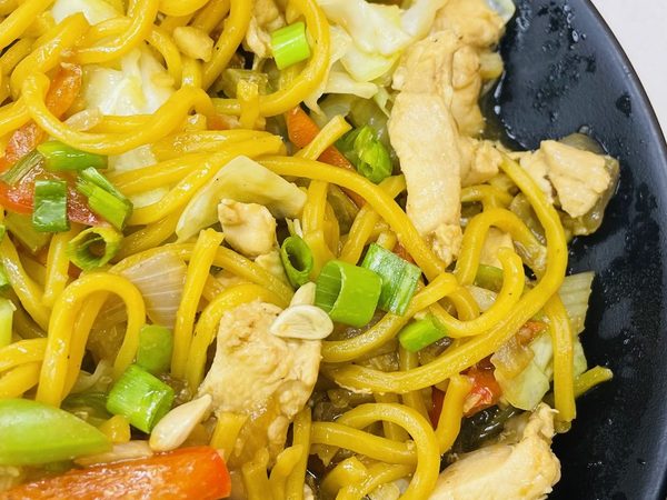 Close-up of chicken pancit canton, yellow egg noodles tossed with cabbage, carrot strips and spring onions on a black plate.
