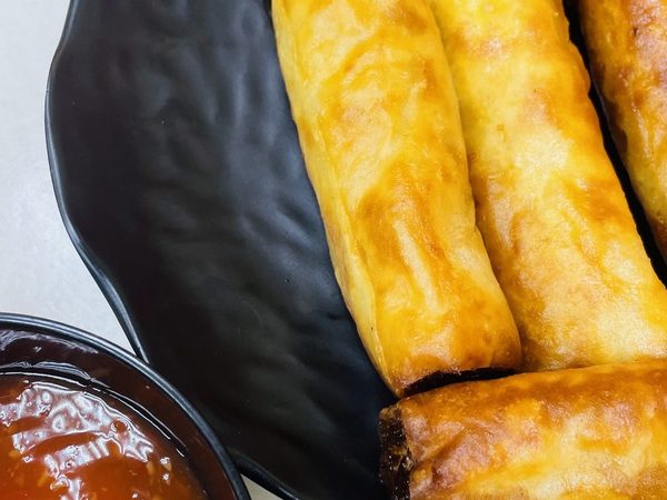 Golden fried lumpia spring rolls stacked on a black petal-shaped plate with a bowl of sweet chilli dipping sauce beside them.