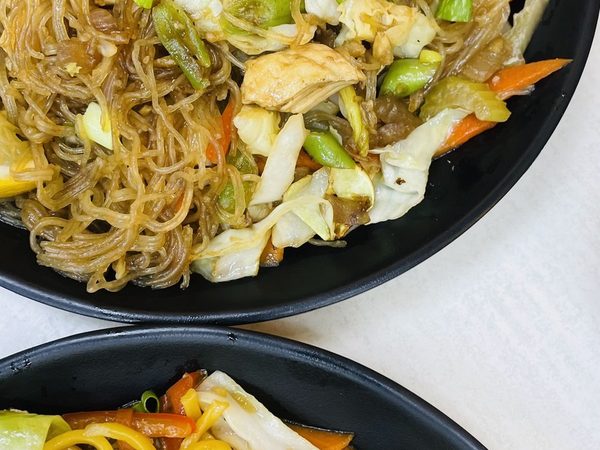 Two black plates side by side holding pancit bihon rice noodles and yellow pancit canton with cabbage, carrot and chicken.