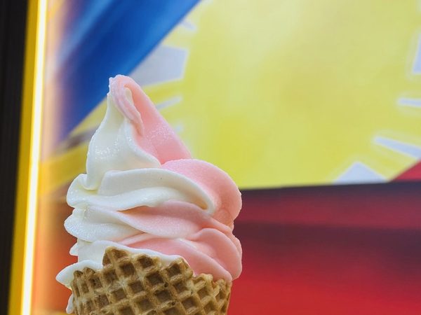 A hand holds a pink and white soft serve swirl in a waffle cone in front of a brightly lit Philippine flag mural.