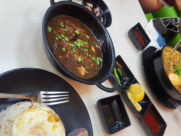 Overhead view of a shared table with crispy chicharon bulaklak, a pot of dark pork dinuguan topped with spring onions and garlic rice with a fried egg.