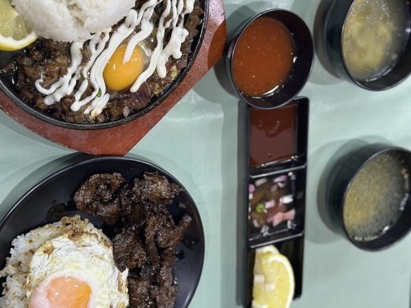 Overhead view of sizzling sisig topped with egg yolk and mayonnaise on a hot plate beside a tapsilog serving, soup bowls and dipping sauces.