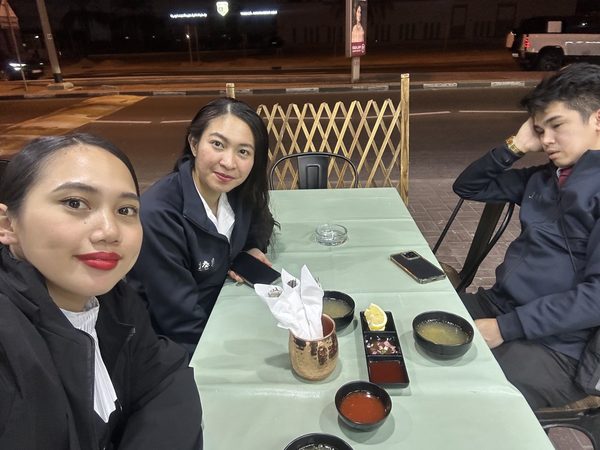 Three friends at a street-side table at night, looking at the camera with soup bowls and dipping sauces set out in front of them.