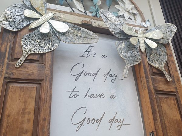 Wall decor of a reclaimed wooden window frame trimmed with large metal butterflies and flowers around a sign reading It's a Good day to have a Good day.