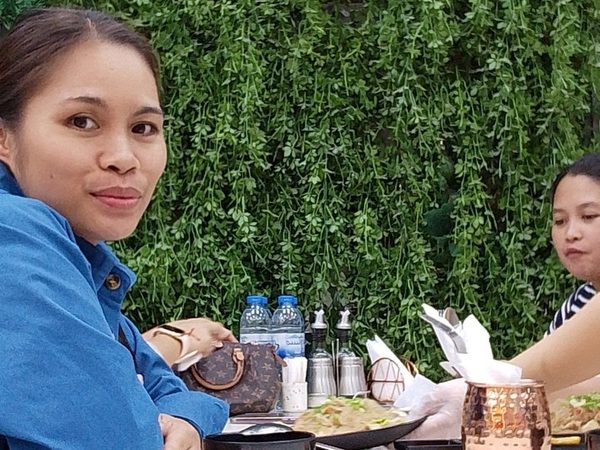 A woman smiles at the camera at a restaurant table in front of a green foliage wall decorated with a bicycle-shaped clock.