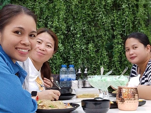 Three women pose together at a table set with pancit and glasses of buko pandan, in front of a green foliage wall with a bicycle-shaped clock.