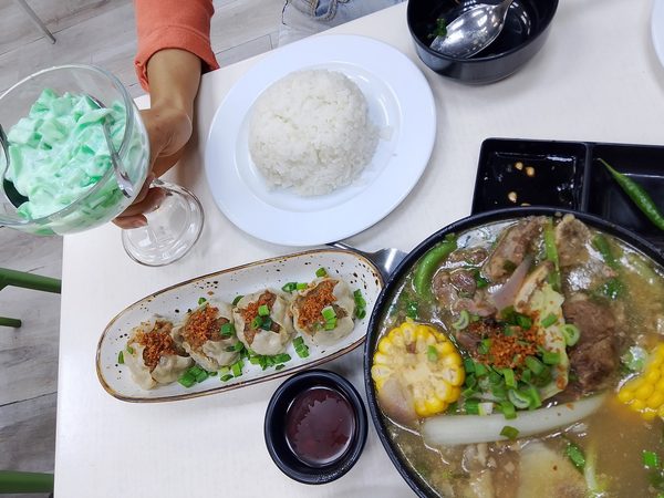 Overhead table spread with a bulalo beef shank hot pot, pork siomai dumplings, steamed rice and a glass of buko pandan dessert.