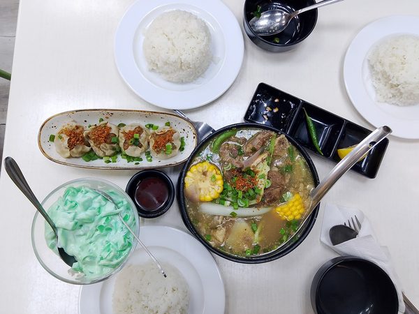 Overhead view of a bulalo hot pot with corn and cabbage beside a plate of siomai, three servings of rice and buko pandan in a glass.