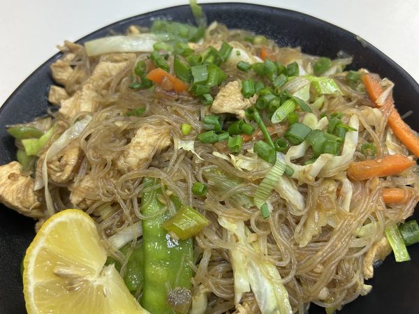 Plate of pancit bihon noodles with chicken, cabbage, carrot and snow peas, topped with spring onion and a lemon wedge.