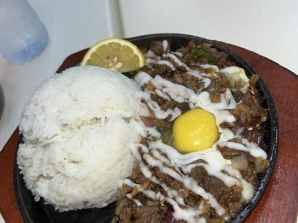 Sizzling sisig on a hot plate with a raw egg yolk and mayonnaise drizzle, served with a mound of steamed rice.