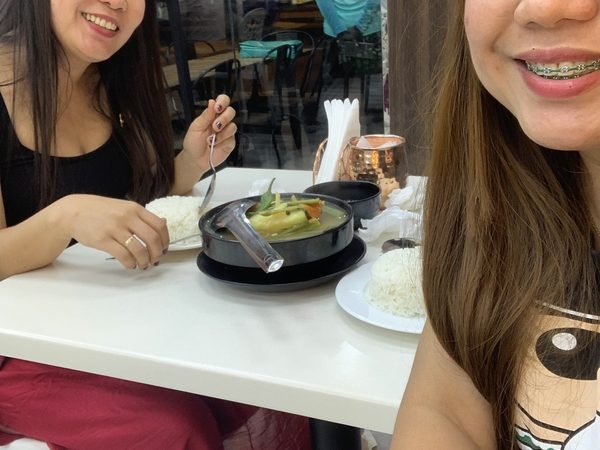 Two women smiling at a dining table over a black pot of sinigang soup and plates of steamed rice.