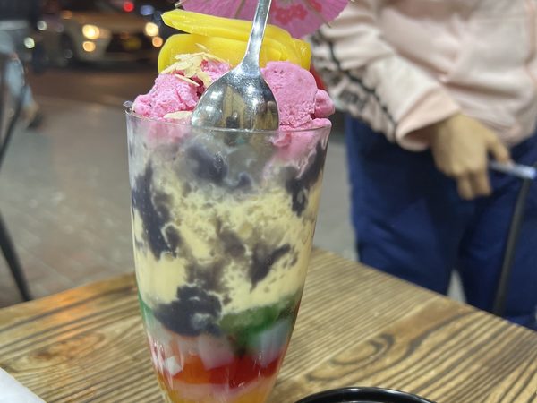 Tall glass of halo halo layered with jellies, leche flan and mango, topped with scoops of ice cream and a paper umbrella.