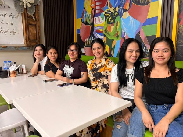 Six women seated together along a green banquette in the dining room in front of a colourful painted mural.