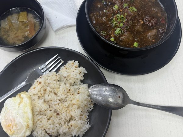 Overhead view of beef pares braised in dark broth with star anise and spring onions, served with garlic fried rice topped with a fried egg and a side bowl of vegetable soup.