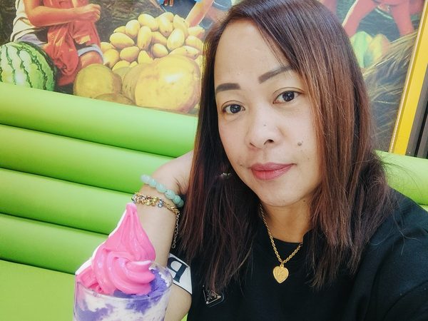 A woman smiling in a selfie beside a tall glass of halo halo topped with pink and white soft serve, under a birthday banner and a Filipino market mural.