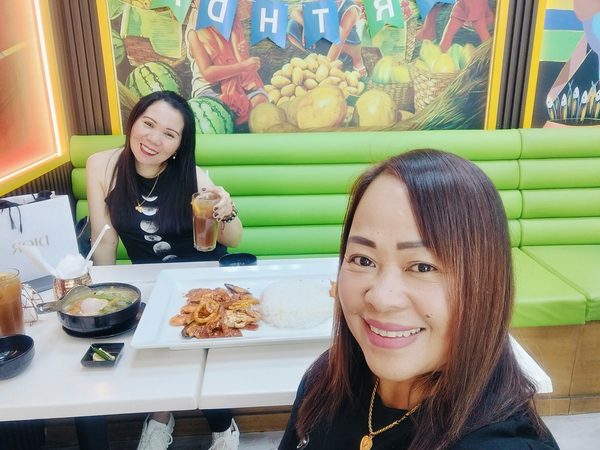 Selfie of two women at a restaurant table with a platter of saucy squid and rice, a bowl of sinigang and iced tea, under a birthday banner and Filipino murals.