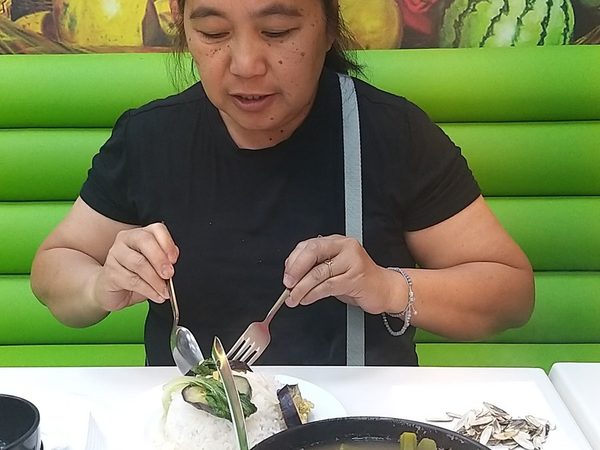 A woman eating fish sinigang from a black bowl of tamarind broth with okra, string beans, radish and green chilli, with a plate of rice beside it.